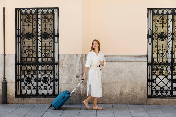 teljes hossza fiatal nő nyári ruha séta poggyász és papír pohár közelében épület Valencia  - Fotó, kép