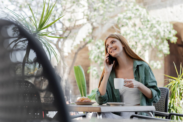 gelukkig vrouw praten op smartphone en het houden van een kopje koffie in de buurt croissant op tafel in cafe terras  - Foto, afbeelding