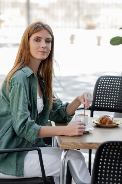 mujer con cabello rojo revolviendo café con cuchara cerca de sabroso croissant en la terraza de la cafetería - Foto, imagen