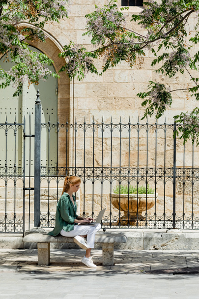 rothaarige Frau sitzt auf Betonbank und benutzt Laptop in der Nähe von geschmiedetem Zaun in Valencia  - Foto, Bild