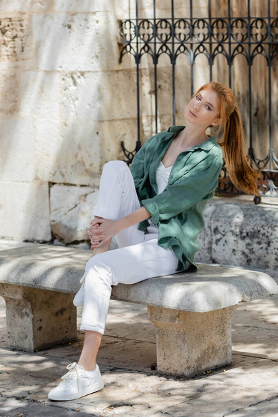 young stylish woman with red hair in green linen shirt sitting on concrete bench near forged fence - Fotó, kép