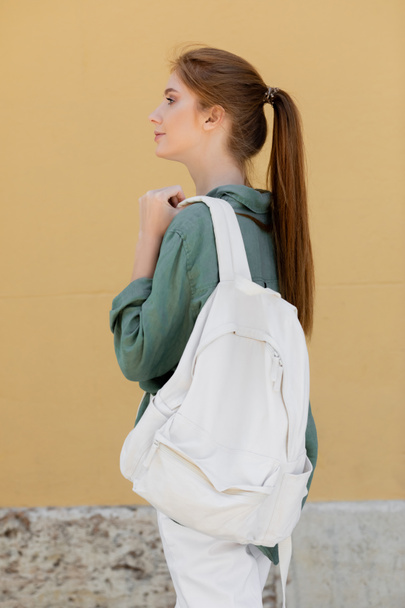 vista laterale della giovane donna con i capelli rossi in camicia di lino verde che tiene lo zaino vicino alla parete beige sulla strada di valencia  - Foto, immagini