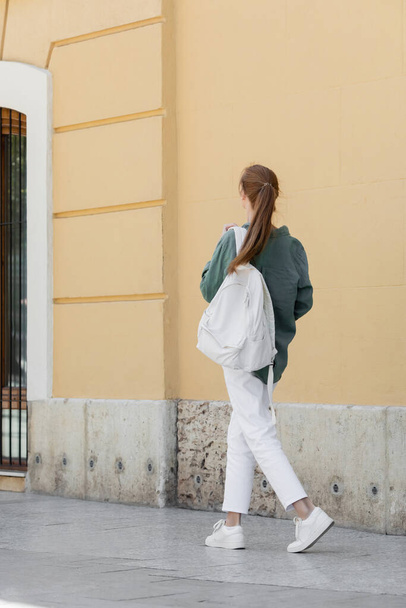 achteraanzicht van roodharige vrouw in groen shirt en witte broek met rugzak en wandelen in de buurt beige muur op straat van valencia  - Foto, afbeelding