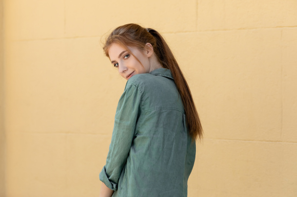молодая женщина в зеленой льняной рубашке позирует возле бежевой стены на улице - Фото, изображение