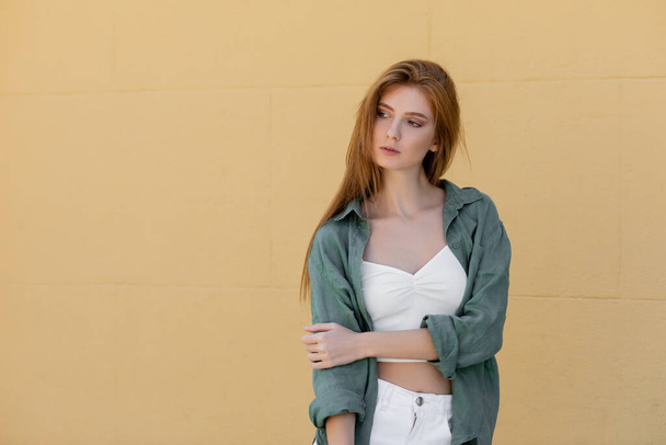 young redhead woman in green linen shirt posing near beige wall on street - Photo, Image