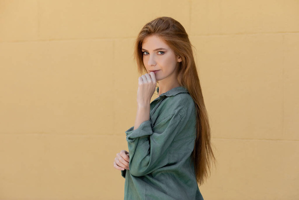 young redhead woman in green linen shirt posing near beige wall  - Fotoğraf, Görsel