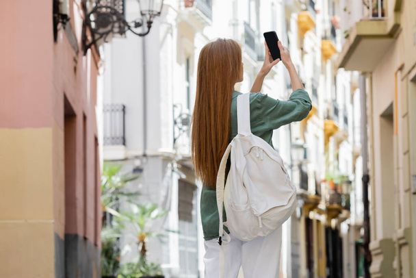 バレンシアの都会の通りでスマートフォンで写真を撮るバックパックを持つ赤毛の女性 - 写真・画像