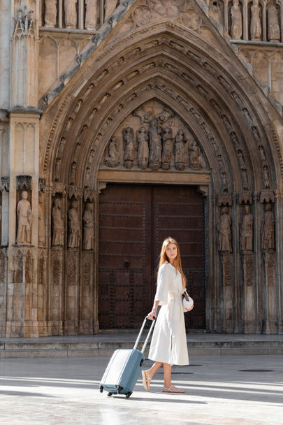 longitud completa de la joven en vestido sosteniendo bolso y de pie con equipaje en valencia - Foto, Imagen