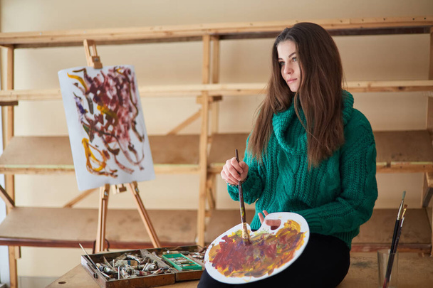 女の子の芸術家、ブルネットは、カラフルな絵を描きます。パレットに絵具を混ぜ、アートワークショップで絵具のプロセスを閉じます。創造的な肯定的な女性画家は、彼女のワークショップで抽象的な絵を描きます. - 写真・画像