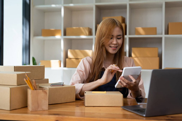 Young woman small business owner online shopping at home. confirm orders from customers with mobile phones preparing package product on background. SME entrepreneur or freelance life style concept - Photo, Image