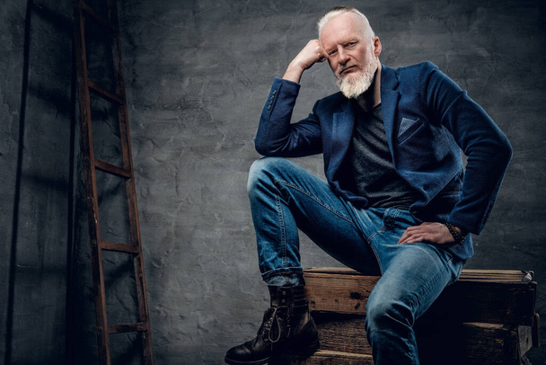 Portrait of gray haired old man with stylish hairstyle sitting on firewoods against dark background. - Valokuva, kuva