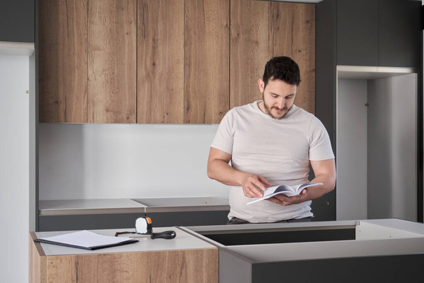 Young argentinian man reading instruction manual before installing furniture on modern new kitchen. - 写真・画像