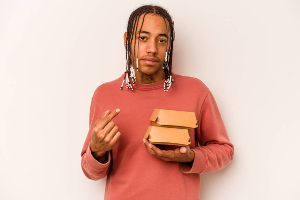 Genç Afrikalı Amerikalı bir adam beyaz arka planda izole edilmiş bir burger tutuyor ve seni işaret ediyor sanki davet ediyormuş gibi.. - Fotoğraf, Görsel