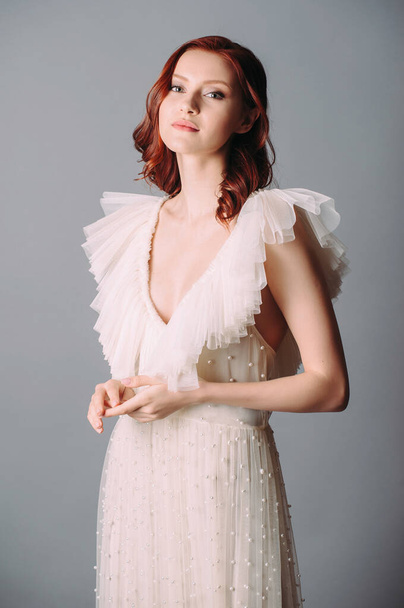 Bohemian wedding, elegant bridal look. Beautiful ginger bride in white wedding dress with sexy decollete and frills on grey background. Candid portrait of a young woman with natural makeup and hairdo. - Фото, зображення