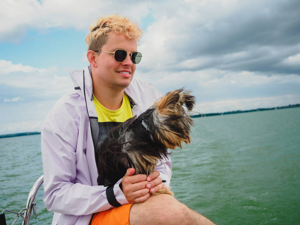 handsome blond young person wear raincoat sitting on a sailing yacht and hold his small dog yorkshire terrier during vacations - Zdjęcie, obraz