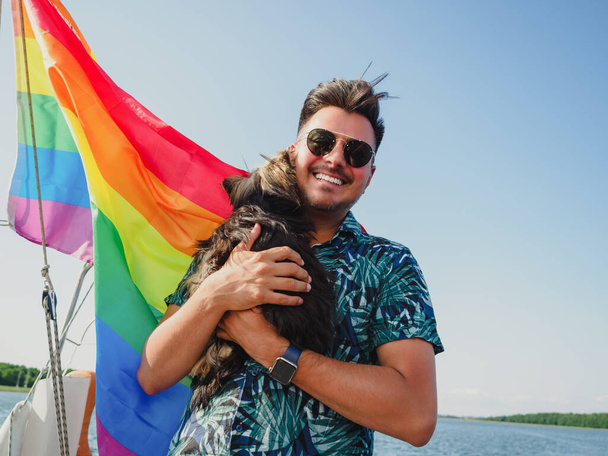 handsome gay person is hugging his small dog yorkshire terrier on a rainbow flag on background - Photo, Image