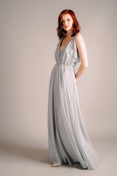Beautiful young woman in dust blue dress. Studio portrait of ginger lady in evening gown dancing on high heel shoes in studio. Bridesmaid's outfit for hen party. - Фото, зображення