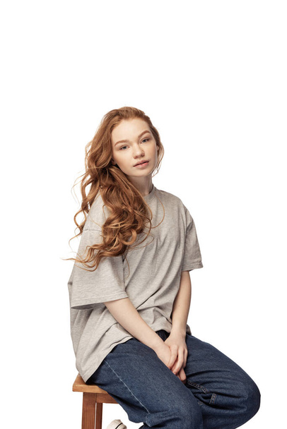 Charming redhead young girl with long curly hair looking at camera with light smile isolated on white background. Female student in casual clothes. Concept of youth, beauty, life. Copy space for ad - Фото, изображение