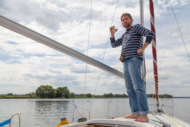 Businessman with a beard in a striped sweater stands on the nose of a white yacht and drinks whiskey from a glas - Foto, Imagem