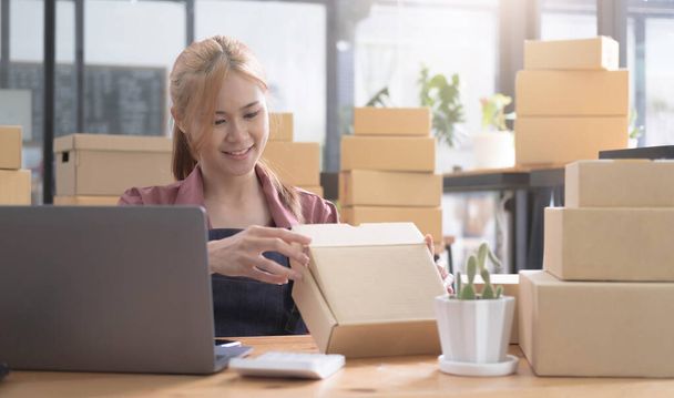 Happy young asian woman startup small business freelance holding parcel box and computer laptop and sitting on chair, Online marketing packing box delivery concep - Photo, Image