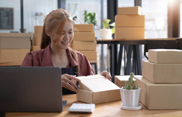 幸せな若いアジアの女性は小さなビジネスフリーランス保持小包ボックスとコンピュータのラップトップを起動し、椅子に座って、オンラインマーケティング梱包箱の配信｜conep - 写真・画像