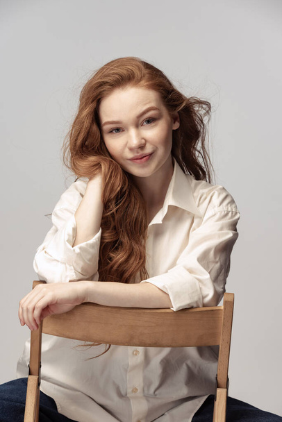 Charming redhead young girl with long curly hair looking at camera with light smile isolated on white background. Female student in casual clothes. Concept of youth, beauty, life. Copy space for ad - Φωτογραφία, εικόνα