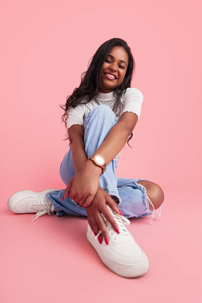 Happy Hispanic teen model in trendy clothes embracing knee and touching white sneaker while sitting against pink background and smiling - Foto, Imagen
