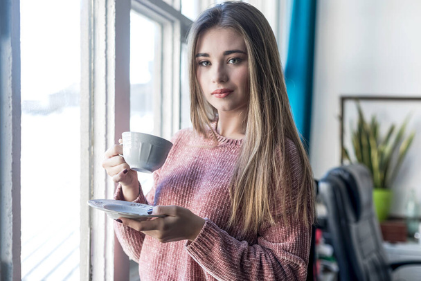 Portrait of a young woman standing next to a window while holding a cup of tea. - Foto, imagen