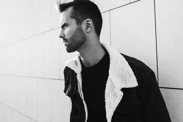 Black and white pensive bearded male in jacket with fur collar frowning and looking away while leaning on tiled wall on city street - 写真・画像
