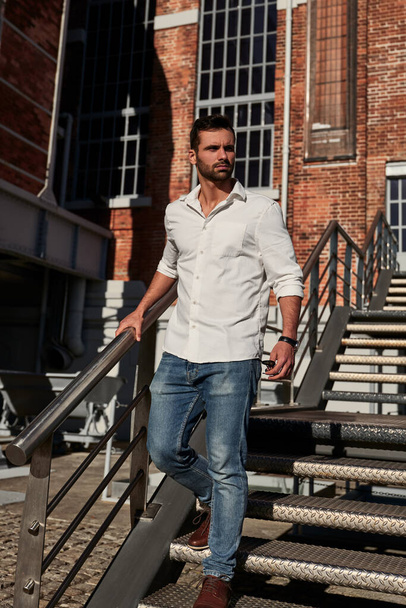 Ethnic bearded male in jeans and white shirt walking down metal stairs and looking away on sunny day outside brick building - Фото, изображение