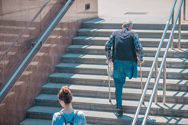 Vanhempi mies, jolla ei ole jalkaa, vamma tai vamma ja kainalosauvat kiipeävät portaita ylös. Julkiset tilat eivät ole vammaisten kannalta valmiita tai epäystävällisiä. - Valokuva, kuva