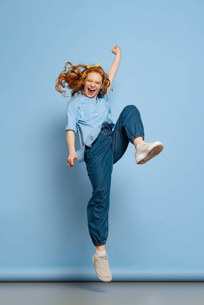 Jumping. Astonished young redhead girl, student posing isolated on blue studio background. Human emotions, facial expression concept. Trendy colors. Youth culture, sales, discount. Copy space for ad - Photo, image