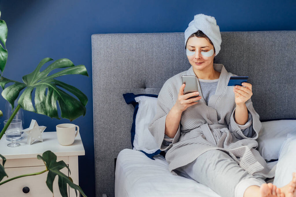 Relaxing woman in pajama and bathrobe with eyes patches and towel on her head sitting in bed while online shopping and make payment online by smartphone and her credit card. Relax and self care - Foto, afbeelding