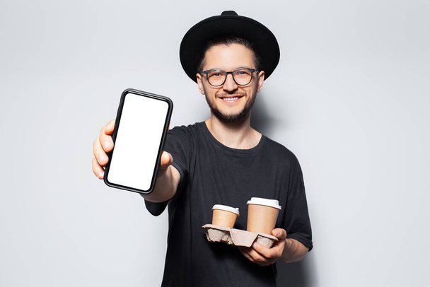 2杯のコーヒーを持っている若い幸せな男は、モックアップ付きのスマートフォンを示し. - 写真・画像