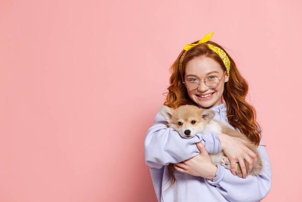 Beloved pet. Cheerful redhead young girl with long curly hair holding cute little puppy of corgi dog isolated on pink background. Care, support and love. Concept of youth, beauty, life. Copy space for - Фото, изображение