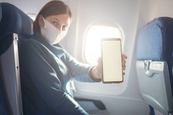 Junge schöne Frau, die während des Fluges am Flugzeugfenster sitzt. neue normale Fahrt nach Covid-19 Pandemiekonzept, zeigt Smartphone-Monitor-Bildschirm, Attrappe - Foto, Bild
