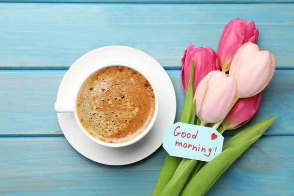 Cup of aromatic coffee, beautiful pink tulips and Good Morning note on light blue wooden table, flat lay - Foto, imagen