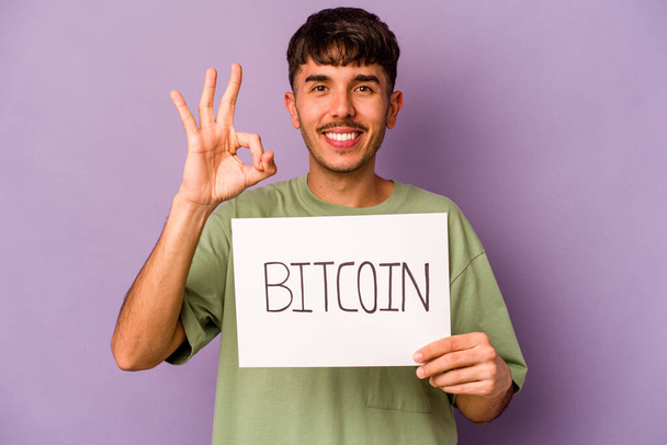 Junge hispanische Mann hält Bitcoin-Plakat isoliert auf lila Hintergrund fröhlich und zuversichtlich zeigt ok Geste. - Foto, Bild