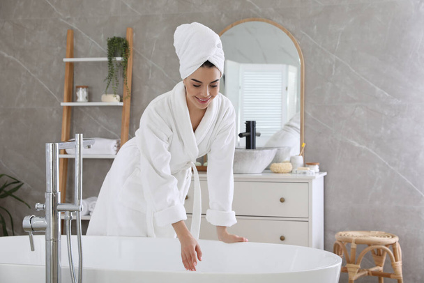 Beautiful young woman in soft white robe near bathtub at home - Foto, Bild