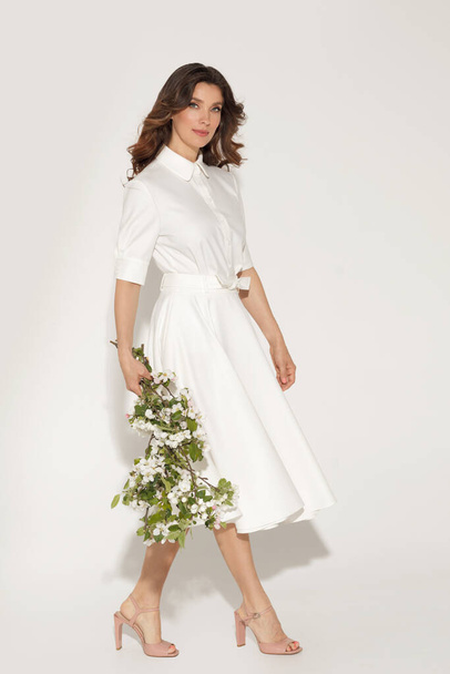波状の髪と白い背景にポーズを自然な新鮮な化粧と白いドレスで自信を持ってブルネットの女性。春や夏のエレガントなファッションコンセプト - 写真・画像