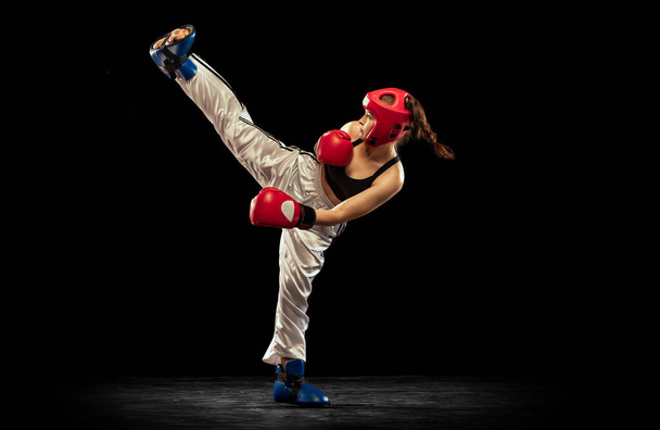 High kick. Attractive female boxer in boxing gloves and helmet training isolated on dark studio background. Sport, competition, hobby, results, success concept. Achievements and challenges - Foto, Imagem