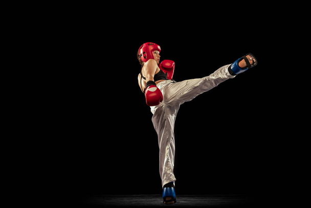 In action. Sportive woman, professional boxer in boxing gloves and helmet training isolated on dark studio background. Sport, competition, hobby, results, success concept. Achievements and challenges - Photo, Image