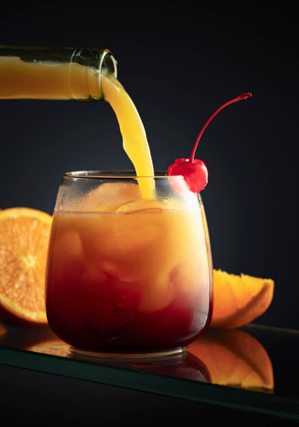 Τεκίλα Sunrise αλκοολούχο κοκτέιλ σε σκούρο φόντο. Χυμός πορτοκαλιού χύνεται σε ένα ποτήρι με τεκίλα, γρεναδίνη και πάγο. - Φωτογραφία, εικόνα