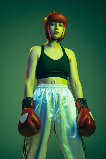 Champion. Professional female boxer in sports protective equipment posing isolated on cyan color studio background in neon. Sport, competition, hobby, results, success concept. Bottom view - Zdjęcie, obraz