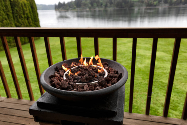Hořící plyn ohniště, přírodní lávové kameny, na venkovní domovské palubě s jezerem a lesy v pozadí  - Fotografie, Obrázek