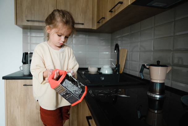 A small child with a rag in his hands stands on a chair near the sink and washes dishes. A little girl helps her mother with cooking and cleaning in the kitchen - Foto, Imagen