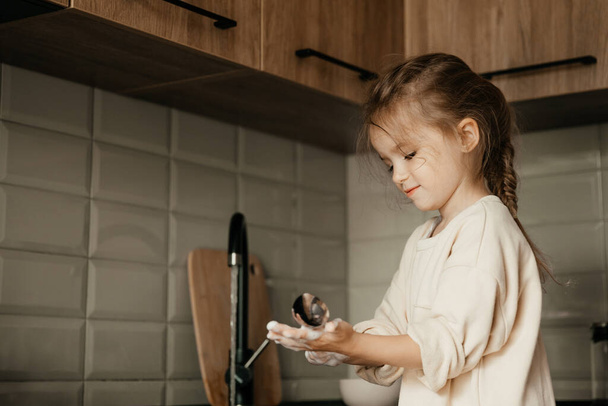A small child with a rag in his hands stands on a chair near the sink and washes dishes. A little girl helps her mother with cooking and cleaning in the kitchen - Foto, Bild