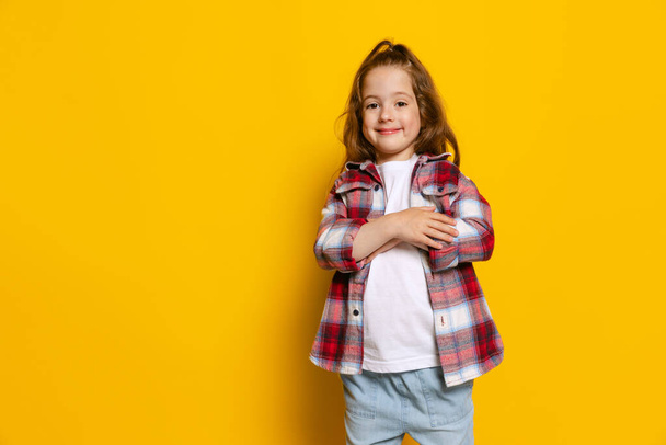 Looks delighted. Happy little girl, kid wearing warm plaid shirt isolated on bright yellow background. Concept of children emotions, fashion, beauty, school and ad concept. Kids fashion, street style - Photo, image