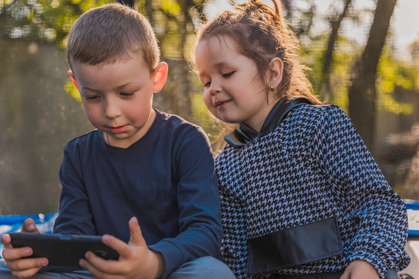 children look at the phone while sitting on a trampoline - Φωτογραφία, εικόνα