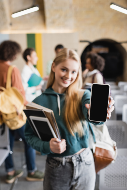 estudiante borroso sosteniendo teléfono inteligente con pantalla en blanco cerca de los estudiantes interracial en el aula - Foto, Imagen